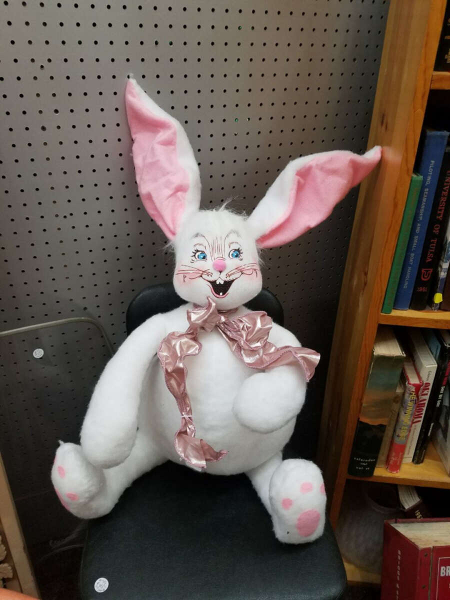 A plush white bunny with large pink ears, blue eyes, and a pink nose sits on a black chair. It has a shiny pink ribbon around its neck and pink spots on its feet. Shelves of books are visible to the right.