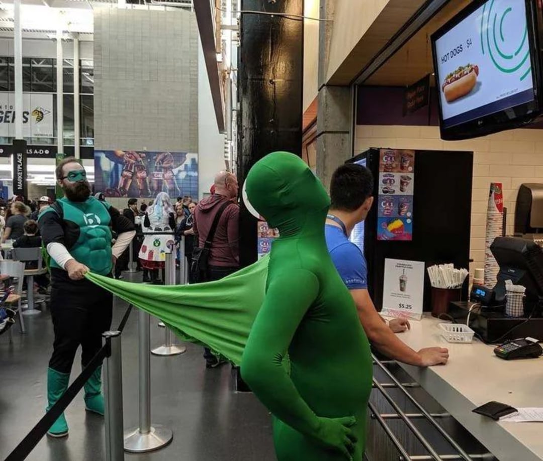 Two people in costumes stand in line at a snack counter. One, dressed as Green Lantern, is stretching the suit of a person in a full green bodysuit. The green-suited person stands at the counter with others around.