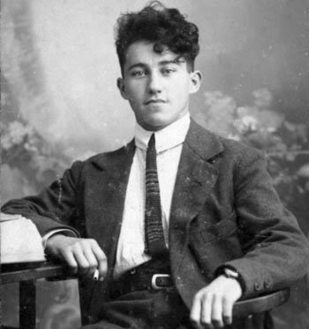 Black-and-white portrait of a young man with curly hair, wearing a suit and tie, sitting at a table with one arm resting on the surface and looking directly at the camera.