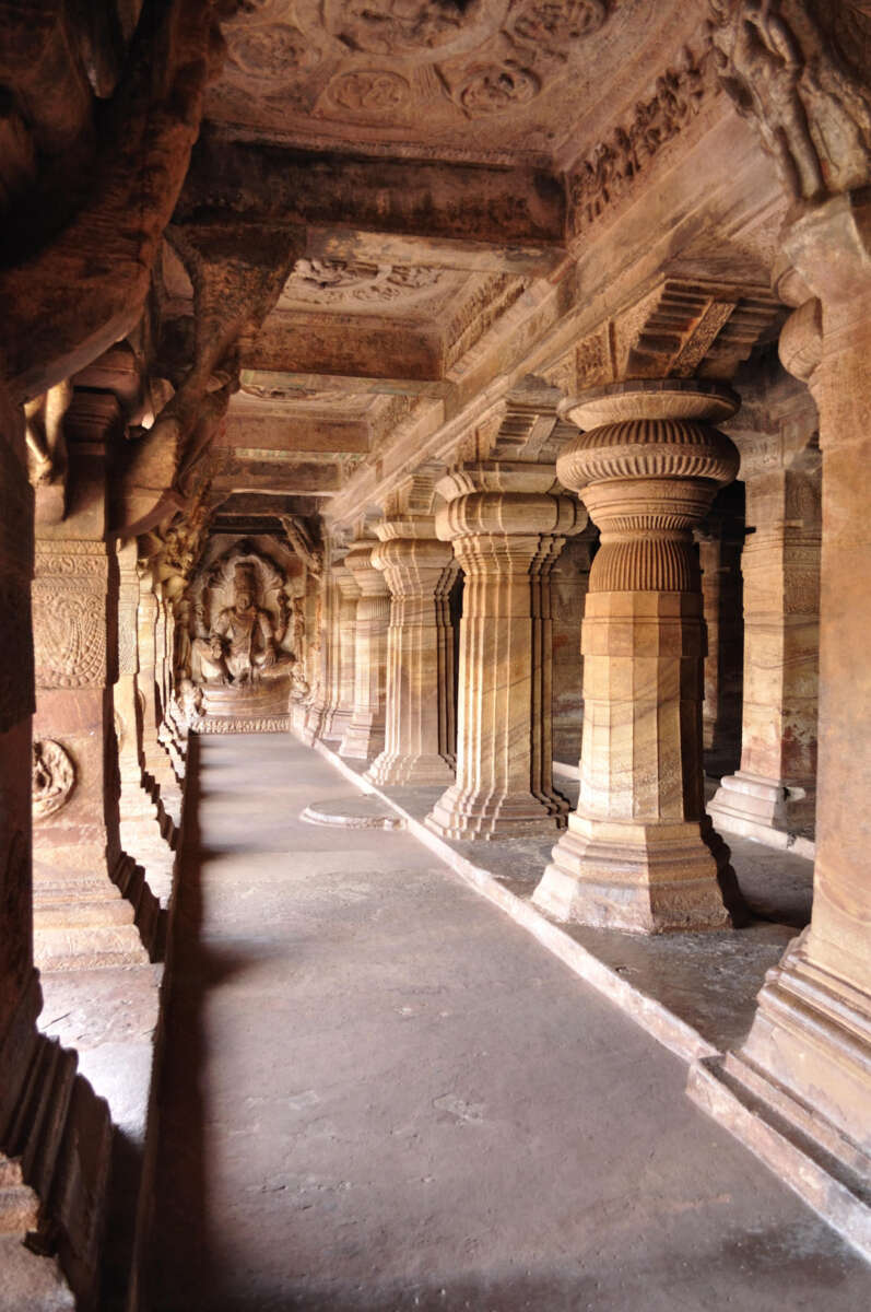 Ancient stone temple corridor with intricately carved columns and ornate ceiling, leading to a detailed statue of a Hindu deity at the end, illuminated by natural light.