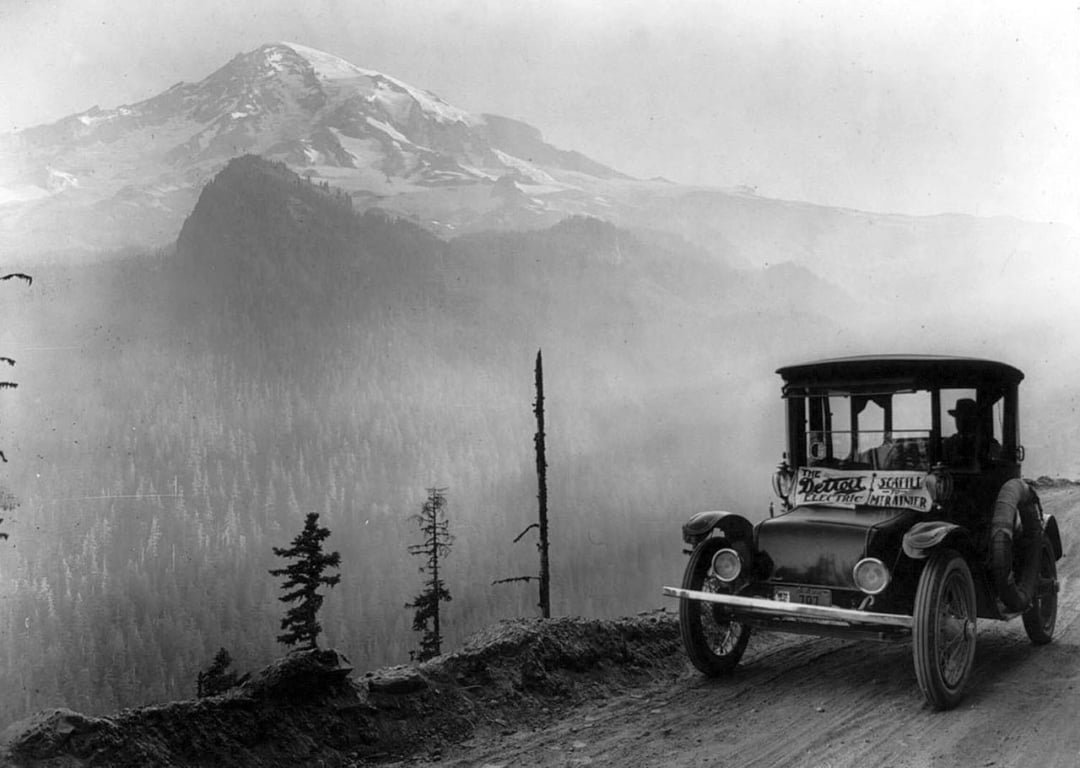A vintage car drives along a narrow, dirt mountain road with a large, snow-capped peak and forested slopes in the background under a hazy sky.