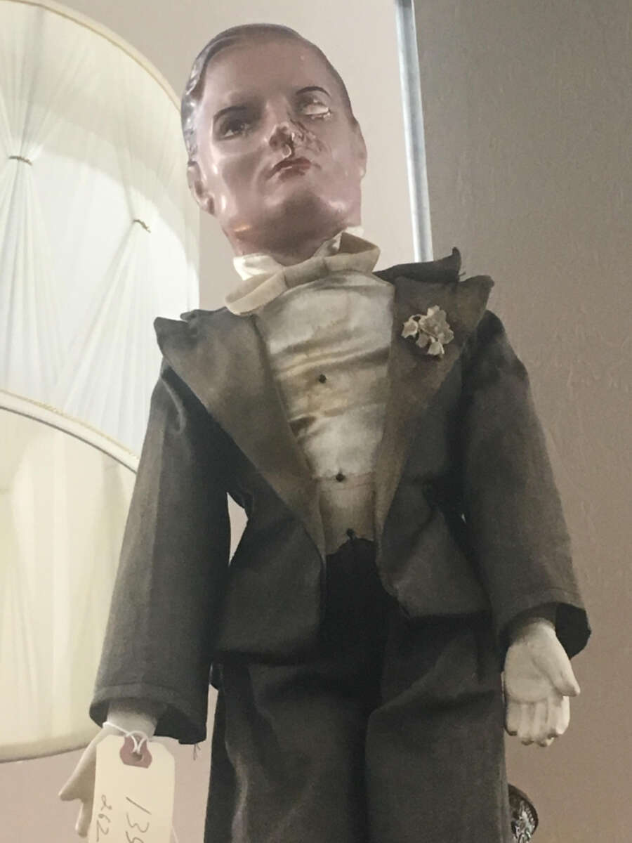 A vintage porcelain doll in a worn gray suit with a white shirt and bow tie, showing a damaged face, stands upright with a price tag on its left wrist. The background features a tall white lampshade and a textured wall.