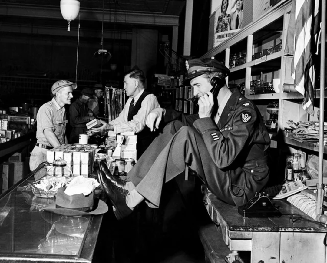 A man in a military uniform sits on a counter, talking on the phone and eating, while two other men stand behind the counter engaged in conversation. Shelves of goods line the background in a busy store setting.
