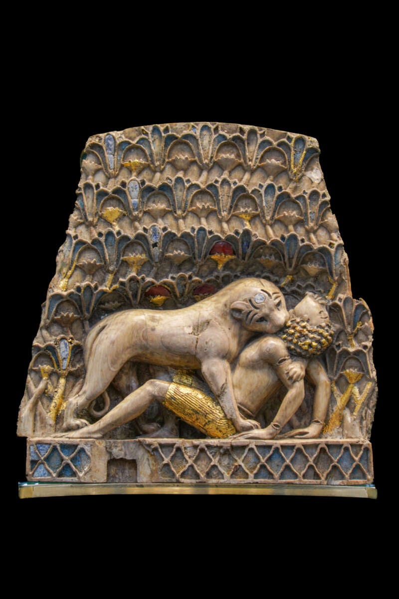 Ivory relief depicting a lion attacking a man, with intricate geometric and floral patterns in gold, blue, and red in the background. The man lies beneath the lion, grasped by its jaws.