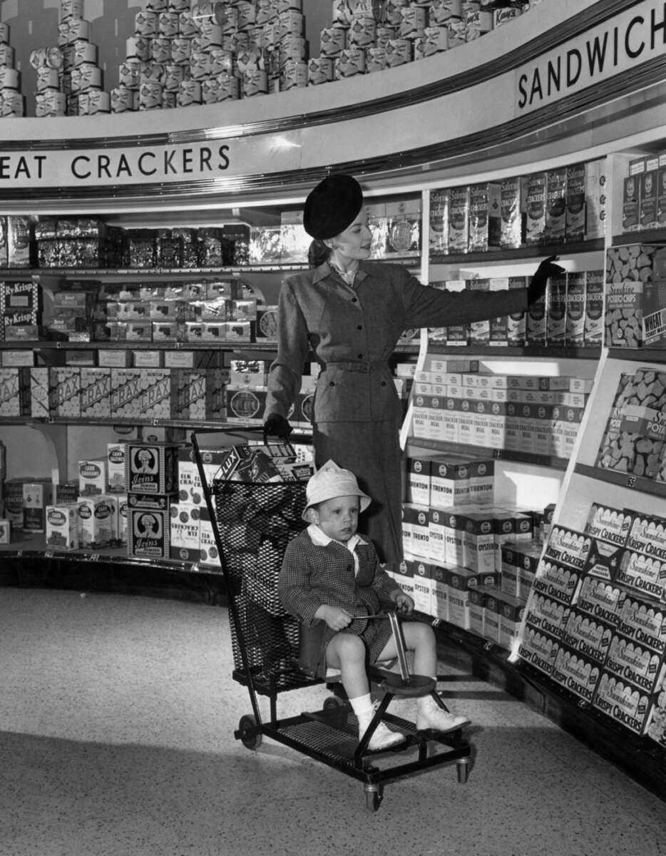 A woman in a 1940s-style outfit shops for crackers in a grocery store, reaching toward shelves. A young child sits in a shopping cart in front of her, looking forward while the woman looks at the products.