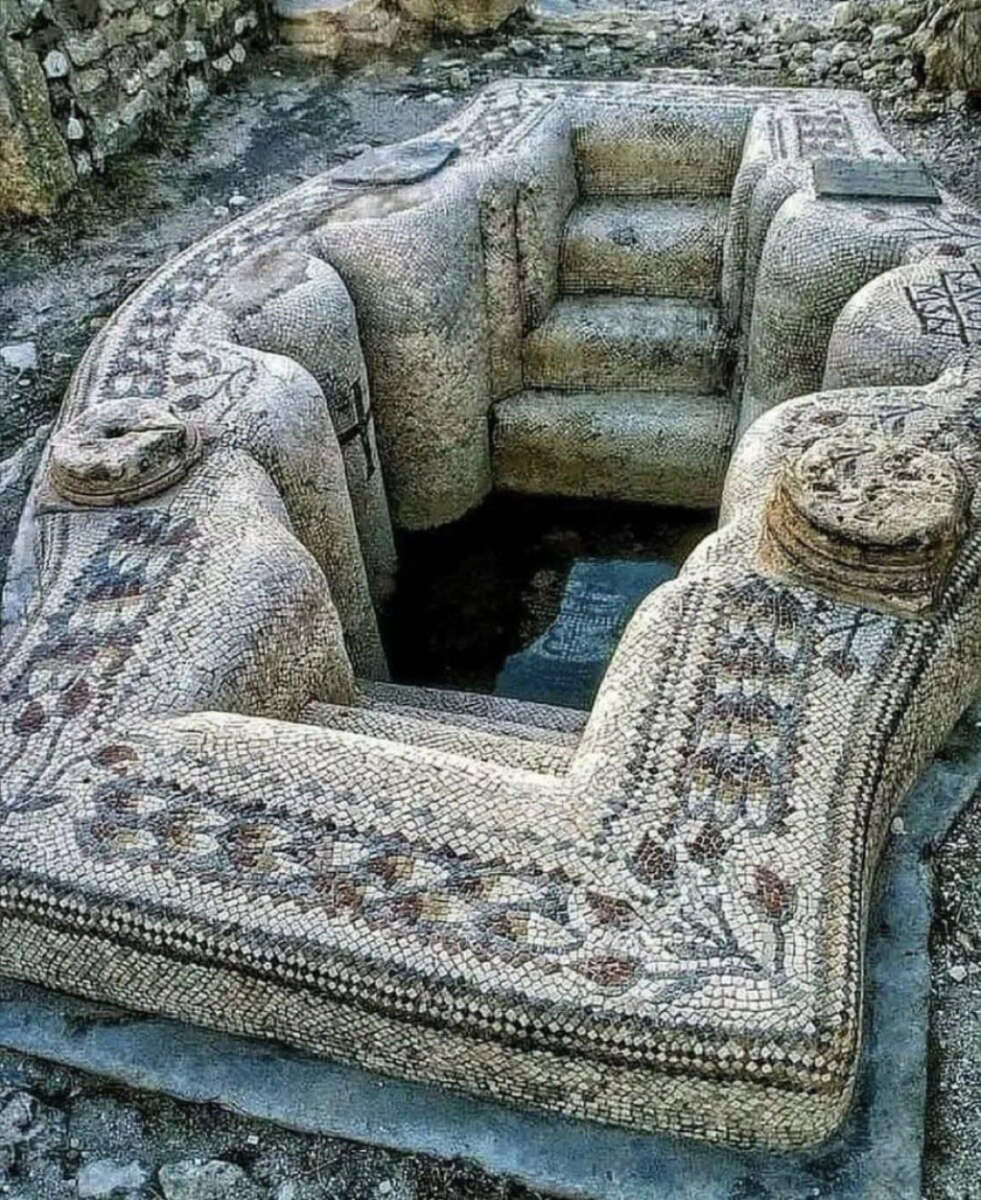 Ancient stone structure shaped like a bathtub or small pool, decorated with detailed mosaic patterns; steps lead down into its center, surrounded by ruins and stones.