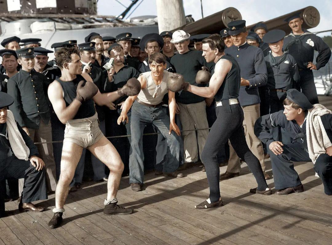 Two men in vintage boxing attire spar on a ship deck, surrounded by sailors in uniforms. One man acts as referee, while the others watch intently, creating a lively scene of camaraderie and competition.