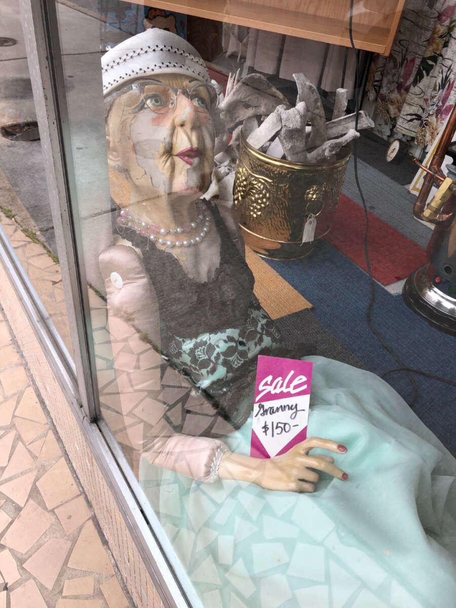 A mannequin dressed as an elderly woman wearing glasses, pearls, and a white hat reclines in a shop window. A sign in front reads "Sale Granny $150–".