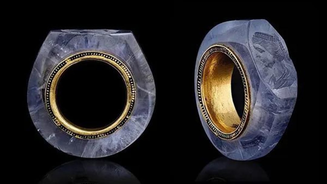 A ring made of translucent blue stone with a gold inner band, shown from the side and at an angle. One side features an engraved profile of a classical-style figure. The ring is set against a black background.