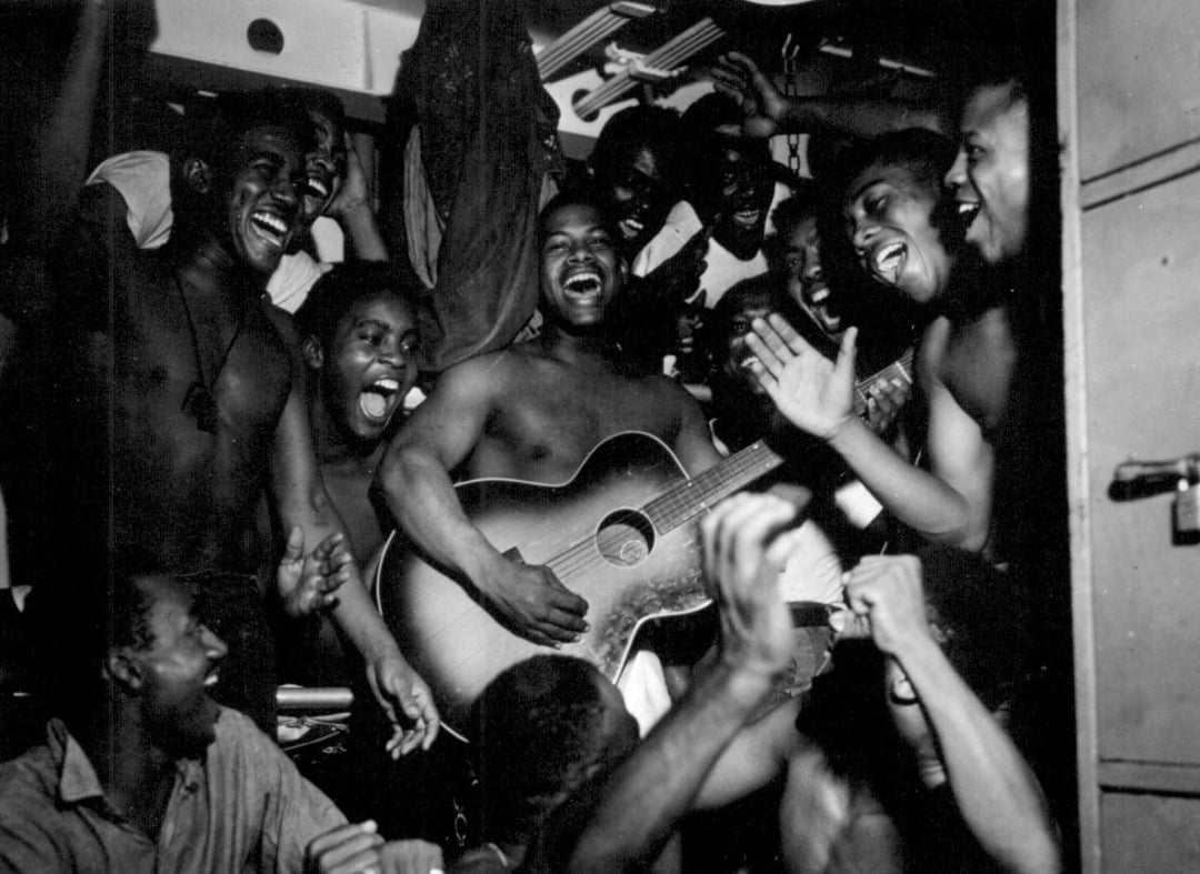 A group of smiling, shirtless men cheer and sing as one man plays an acoustic guitar, creating a joyful, energetic atmosphere in a crowded, dimly lit space.
