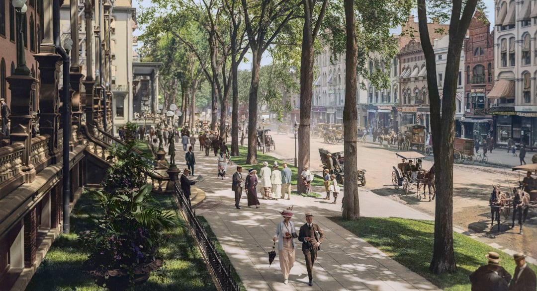 A lively city street scene from the early 1900s shows people walking along a tree-lined sidewalk, vintage carriages, and elegant buildings, with shops and activity on both sides of the street.