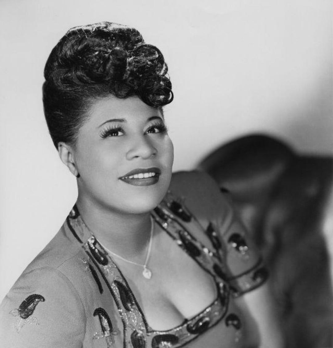 Black-and-white portrait of a woman smiling, wearing a patterned dress and a necklace, with her hair elegantly styled in an updo. She is looking slightly upward with a soft background behind her.