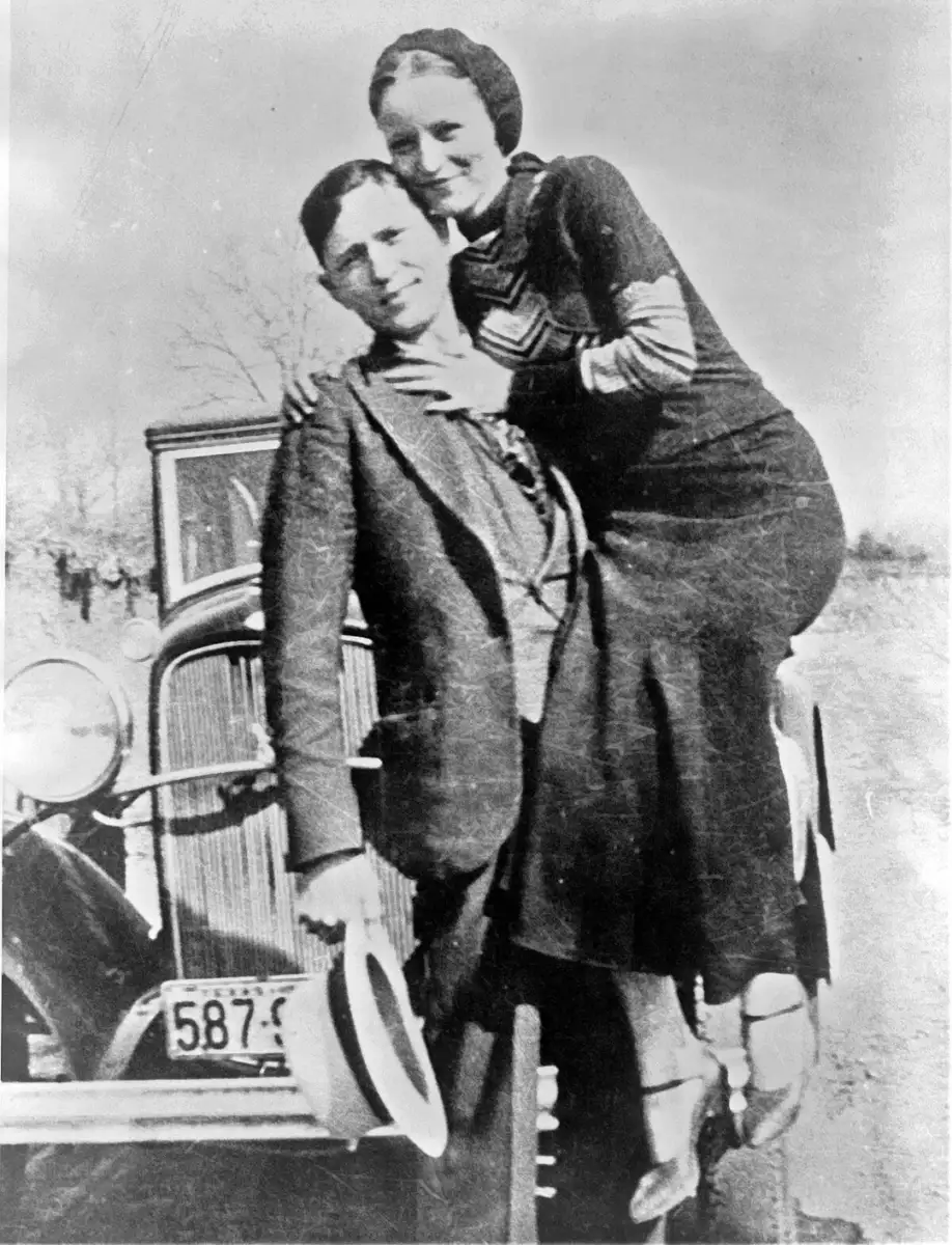 A black-and-white photo showing a man in a suit and hat holding a smiling woman in a dress. They are posed in front of an old car with a visible license plate. The woman is perched on the car's bumper.