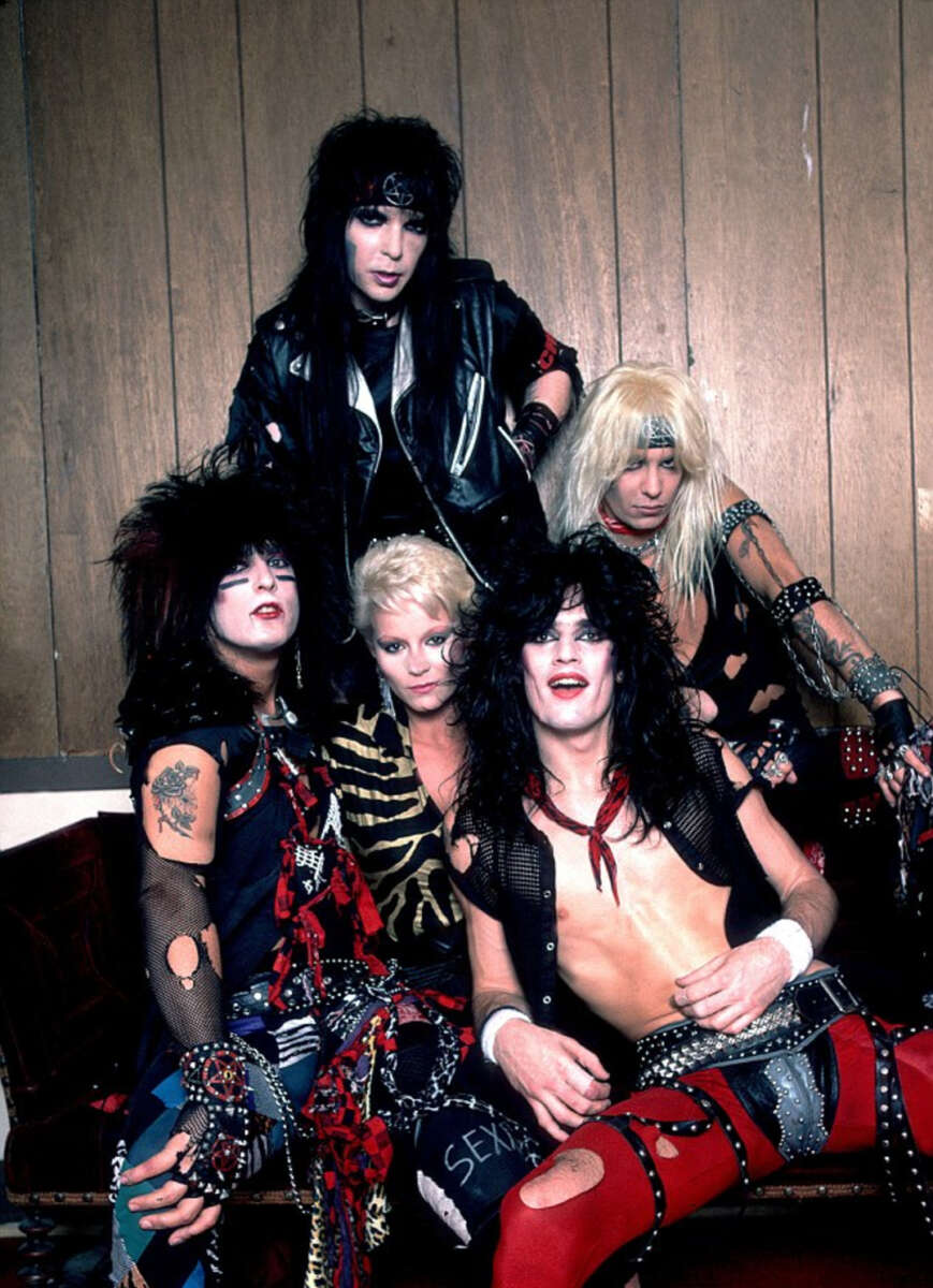 Five people with big, teased hair and glam-rock outfits pose together in front of wood paneling, featuring leather, animal prints, ripped clothing, and bold accessories, evoking 1980s rock band style.