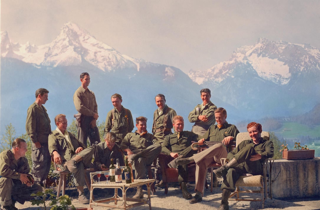 A group of soldiers in green uniforms relax outdoors with drinks, seated and standing on a patio with mountains and snow-covered peaks in the background under a clear sky.