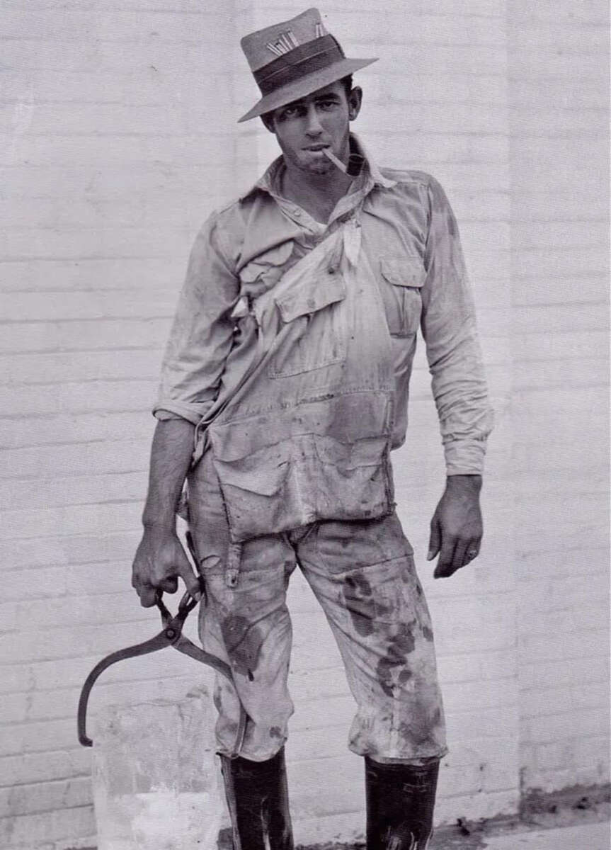 A man wearing a hat, work shirt, and boots stands against a white brick wall, holding a large block of ice with ice tongs. His clothes are dirty and crumpled, and he has a cigarette in his mouth.