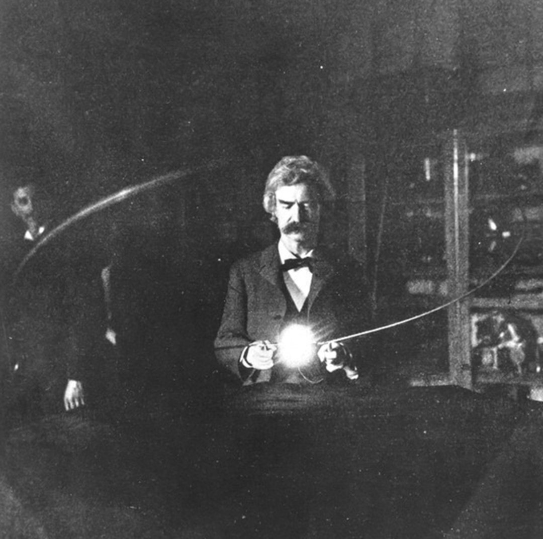 A man in a suit holds a glowing light source, possibly an electrical device, in a dimly lit room with laboratory equipment visible in the background.