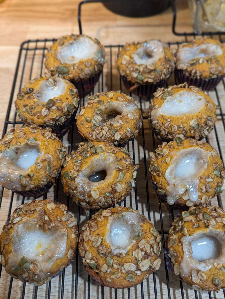 Twelve pumpkin muffins topped with oats and seeds, drizzled with white icing, are cooling on a black wire rack set on a wooden countertop.