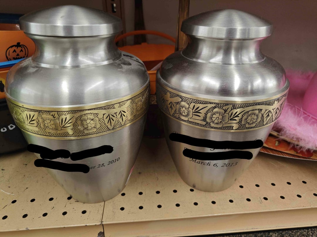 Two silver urns with gold floral bands sit on a store shelf. Each has a blacked-out name and visible dates: "October 28, 2010" on the left urn and "March 6, 2013" on the right.