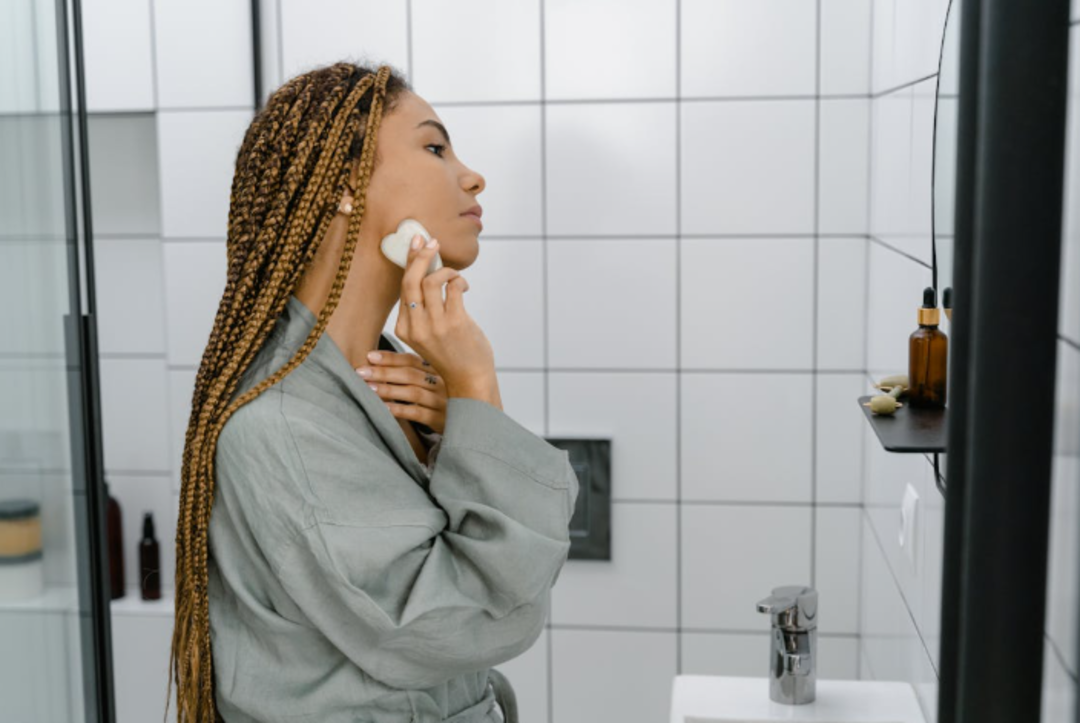 A woman with braided hair wearing a gray robe applies skincare product to her face with a cotton pad in a modern, tiled bathroom. Skincare bottles are visible on a shelf beside her.