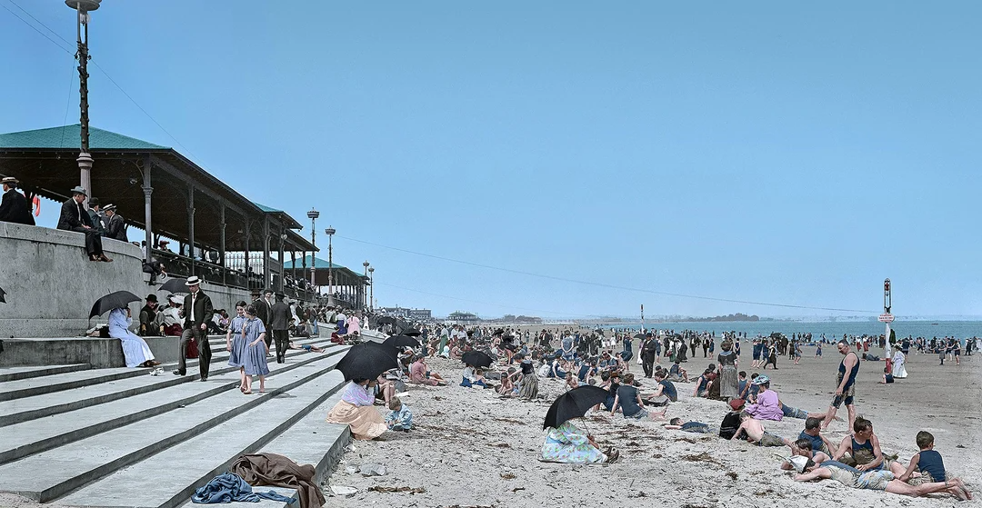 A crowded beach scene with people relaxing on the sand and sitting on concrete steps. Many wear early 20th-century clothing, some with umbrellas for shade, while others wade or walk near the shoreline under a clear blue sky.