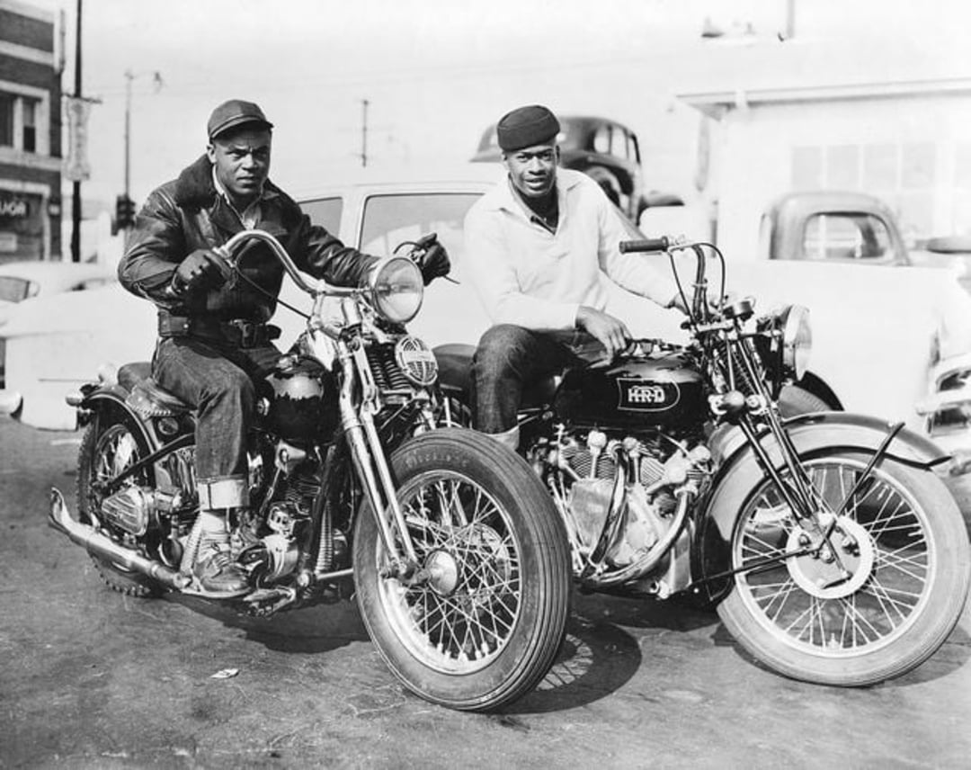 Two men sit side by side on classic motorcycles, wearing jackets and hats, in front of parked cars and buildings, in a black and white vintage street scene.