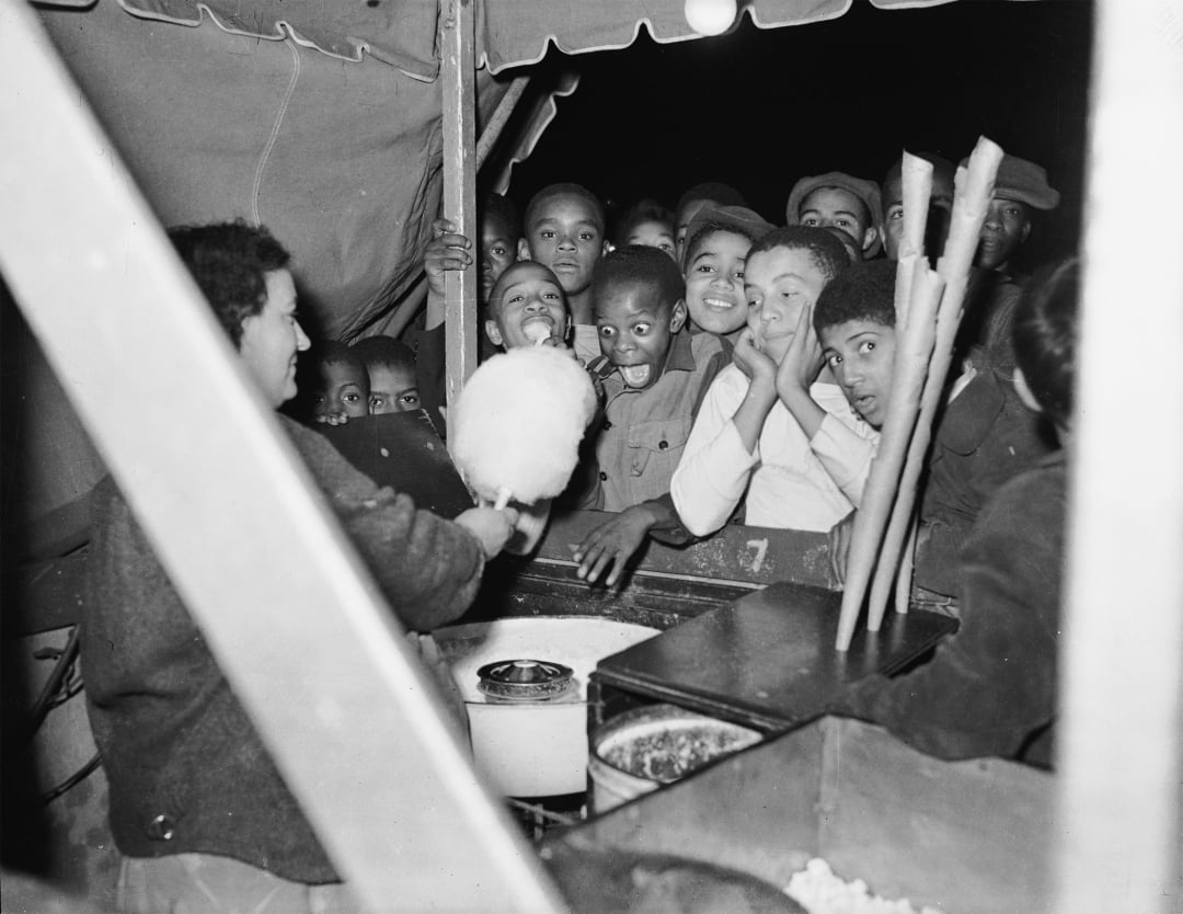 A group of excited children eagerly watch a vendor make cotton candy at an outdoor stand at night, their faces full of anticipation and delight.