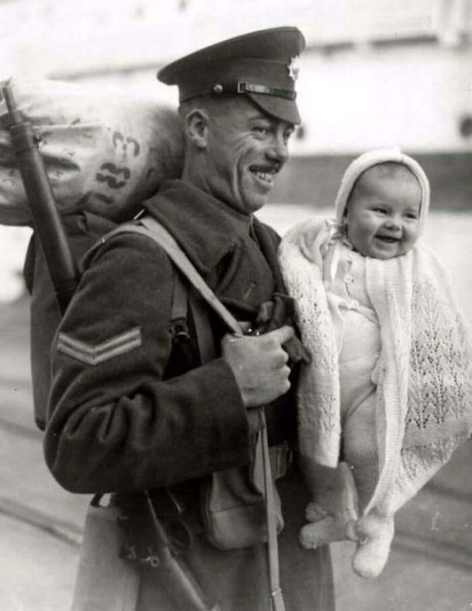 A smiling soldier in uniform carries a large sack on his back and holds a laughing baby wrapped in a knitted blanket, both looking happy.