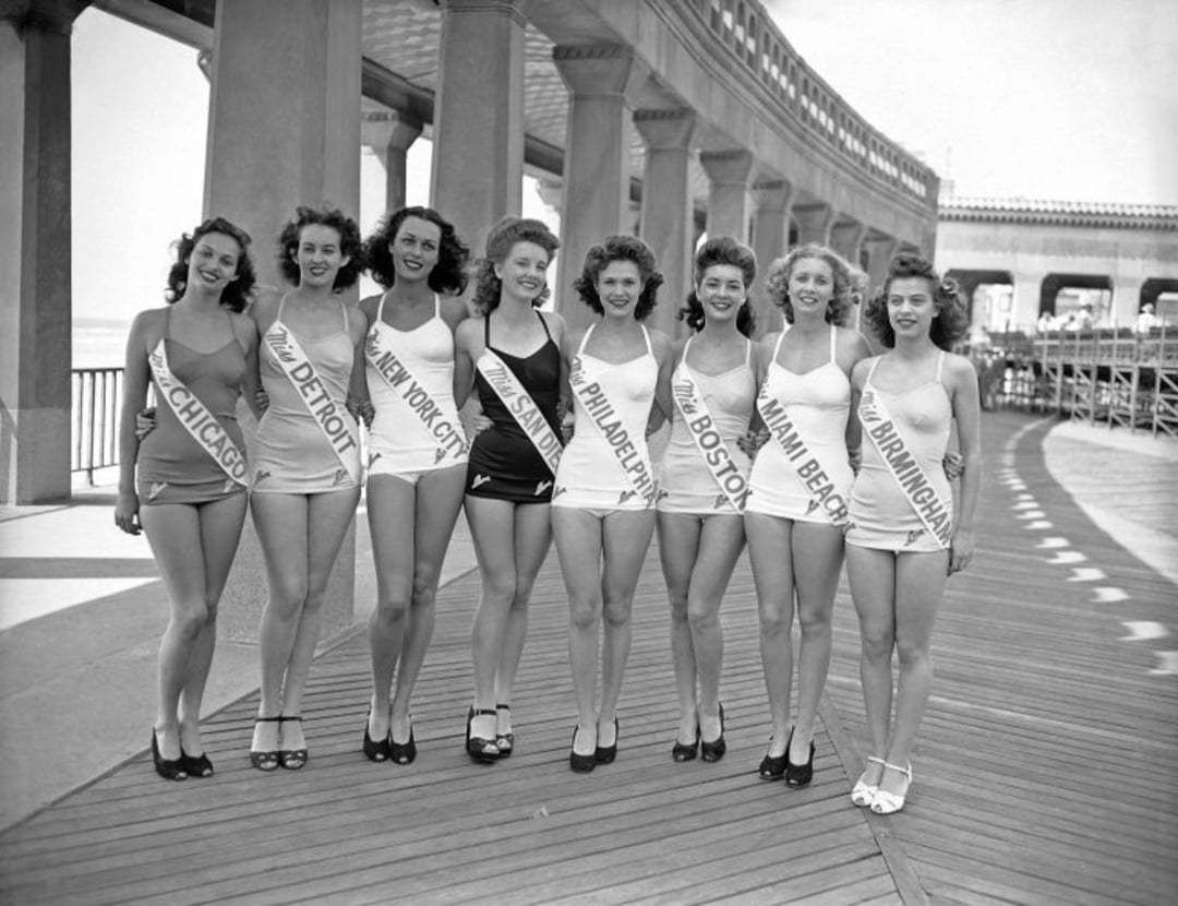 Eight women in swimsuits and high heels stand arm-in-arm on a boardwalk, each wearing a sash with a city name, such as Chicago, Detroit, and New York City. A colonnade and railings are visible in the background.