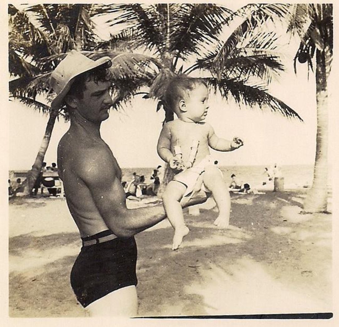 A man in swim trunks and a hat holds a baby on a sandy beach with palm trees, while people relax in the background near the shoreline. The photo is in black and white.