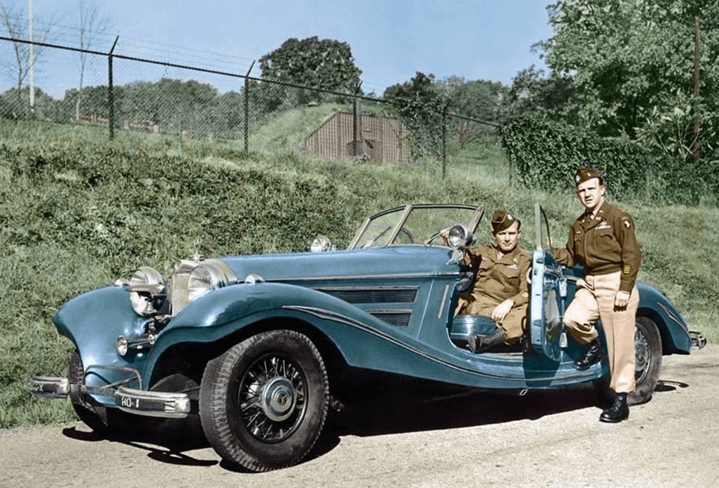 Two men in World War II-era military uniforms pose with a vintage blue convertible car parked on a road, with greenery and a fence in the background. One man is seated in the car, the other stands beside it.