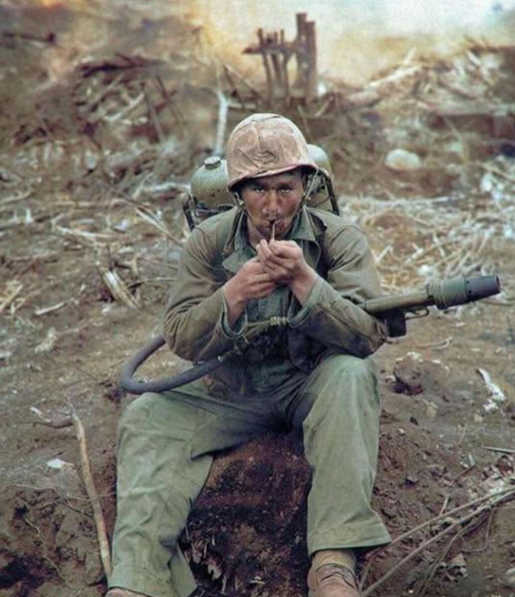 A soldier in a helmet and uniform sits on the ground amid debris, lighting a cigarette. He carries a flamethrower on his back, and the background shows a war-torn landscape.