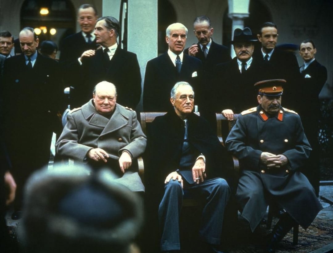 Three men sit on a bench in front of a group of standing men in suits and uniforms. The atmosphere appears formal and historical. The seated men wear heavy coats, with one in a military uniform.