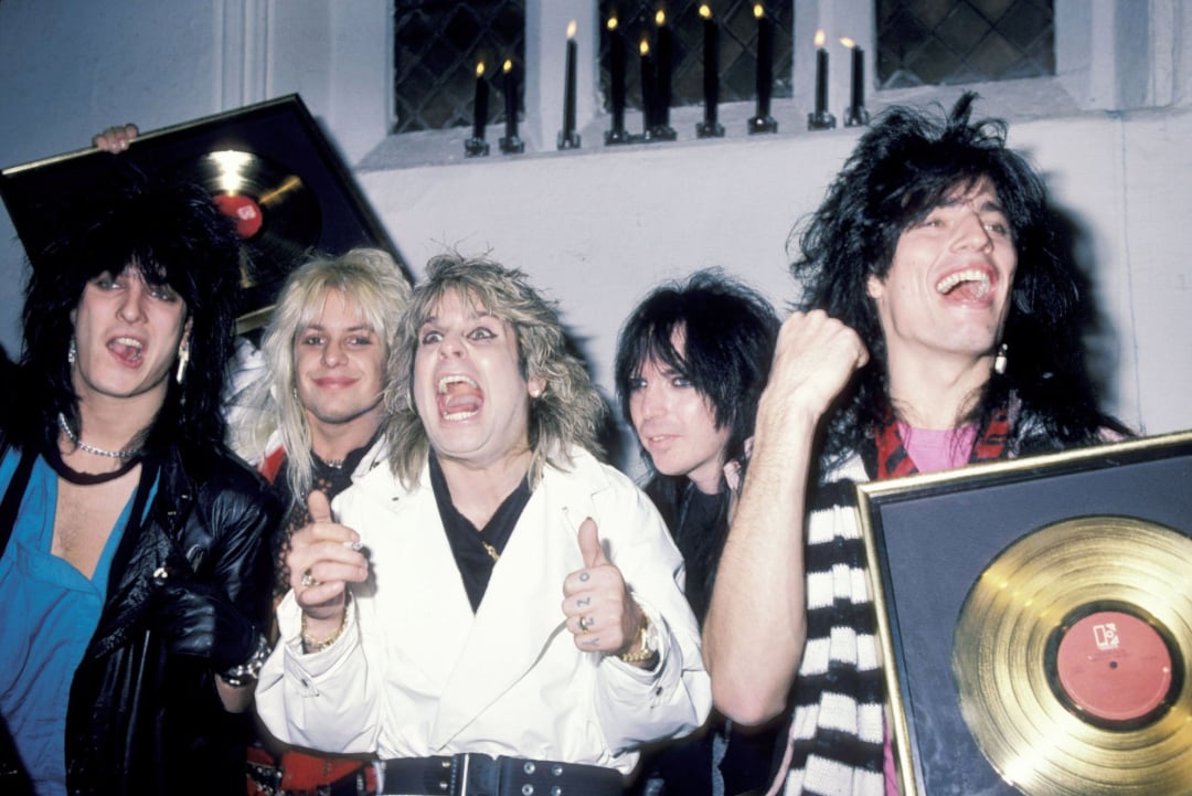 Five excited men with big hair and rock-style clothing hold gold records and celebrate in front of a white wall with black candles. They are smiling and cheering, appearing energetic and happy.