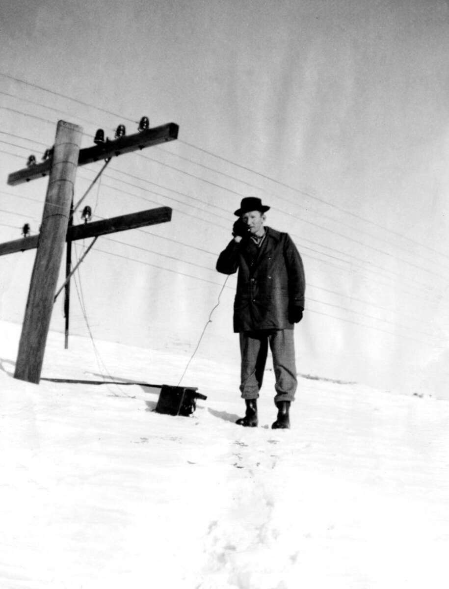 A man in a coat and hat stands in the snow, using a telephone connected to a pole with overhead wires on a wintry landscape under a clear sky.
