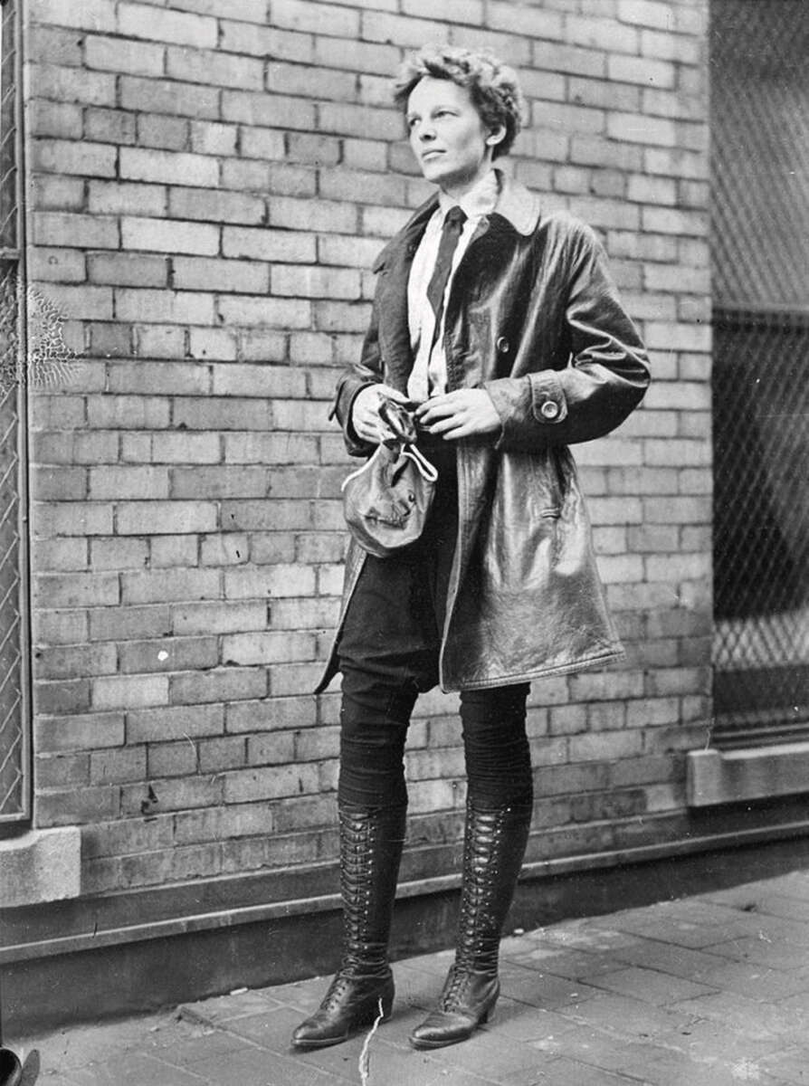 A woman in a leather coat, tie, and tall lace-up boots stands in front of a brick wall, holding a small bag and looking to the side.
