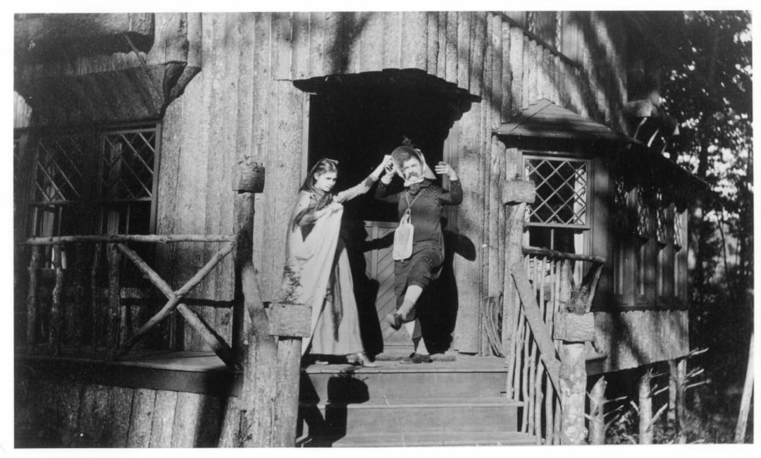 Two people in old-fashioned clothing stand on the steps of a rustic wooden cabin, holding hands and raising one arm in the air as if dancing or celebrating. The cabin has a porch with railings and decorative window panes.