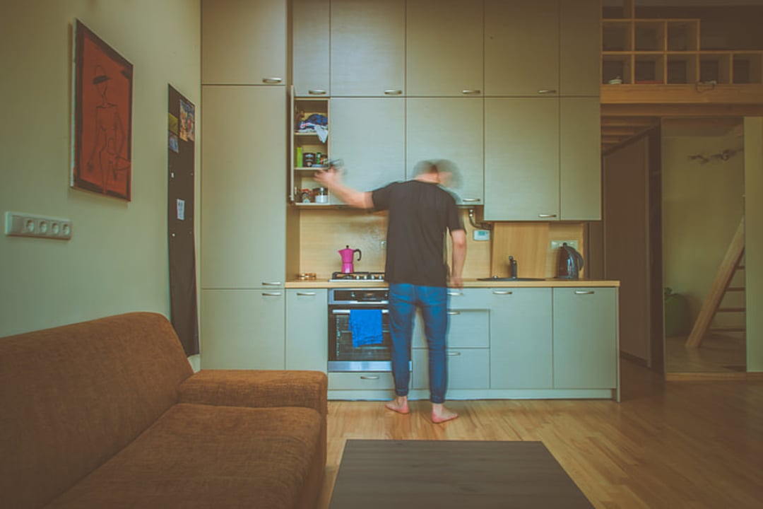 A barefoot person in motion stands in a modern kitchen, reaching toward an overhead shelf. The room features light wood cabinets, a brown sofa, and a hardwood floor.