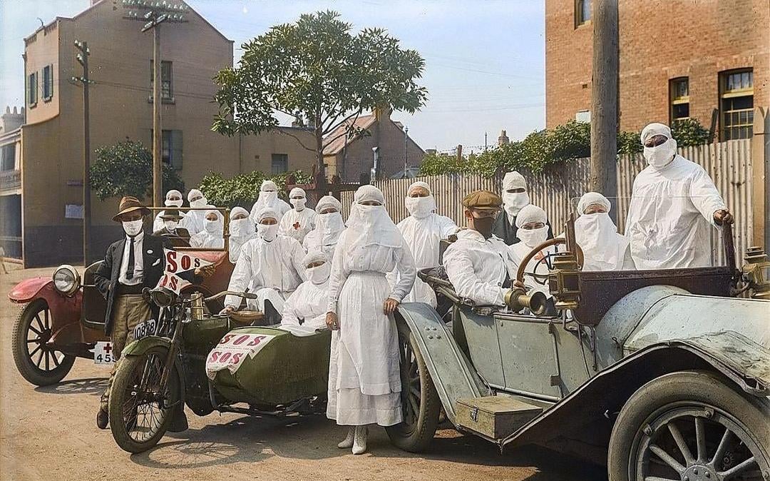 A group of people in white protective clothing and face masks stand and sit around early 20th-century vehicles, including cars and a motorcycle with "SOS" signs, on a city street in front of brick buildings.