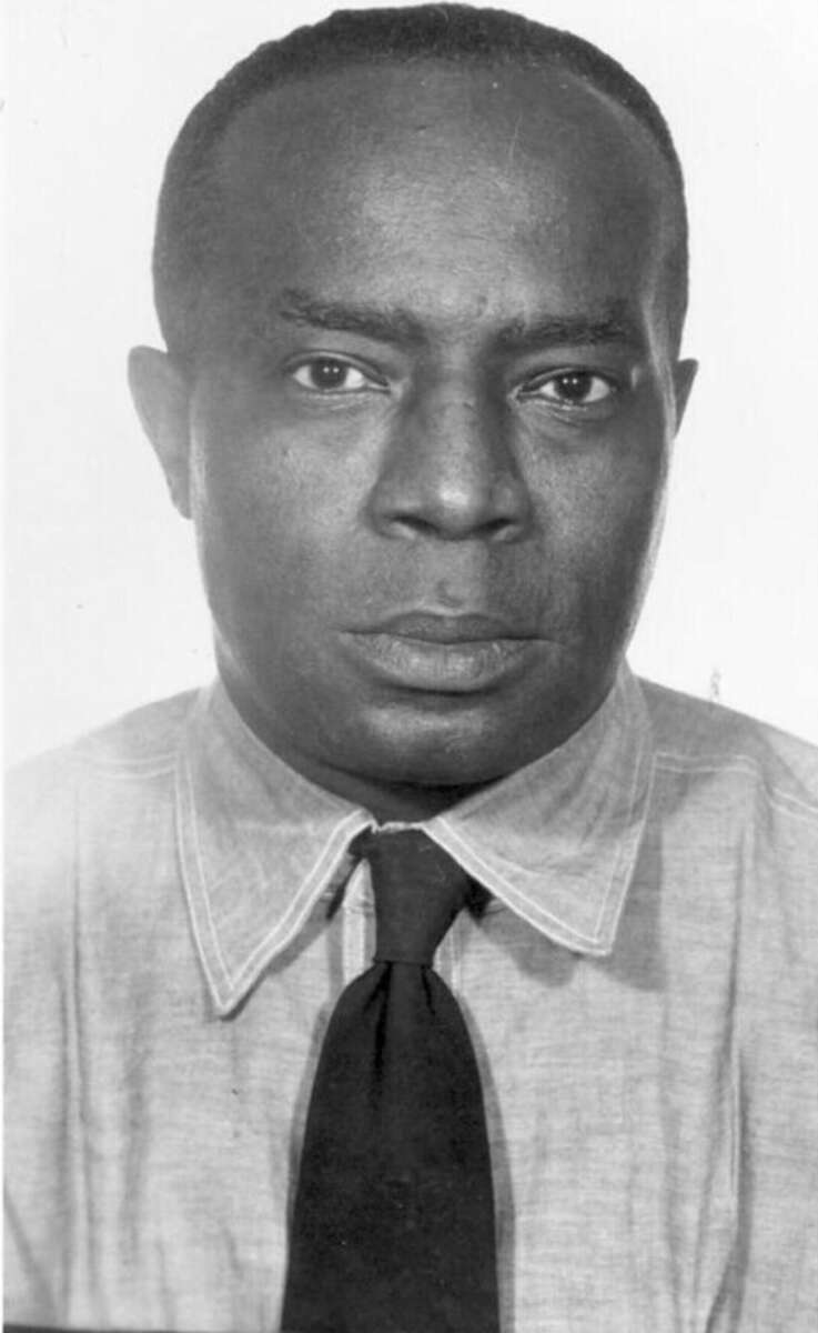 Black-and-white portrait of a man wearing a light-colored button-up shirt and a dark tie, looking directly at the camera with a neutral expression.