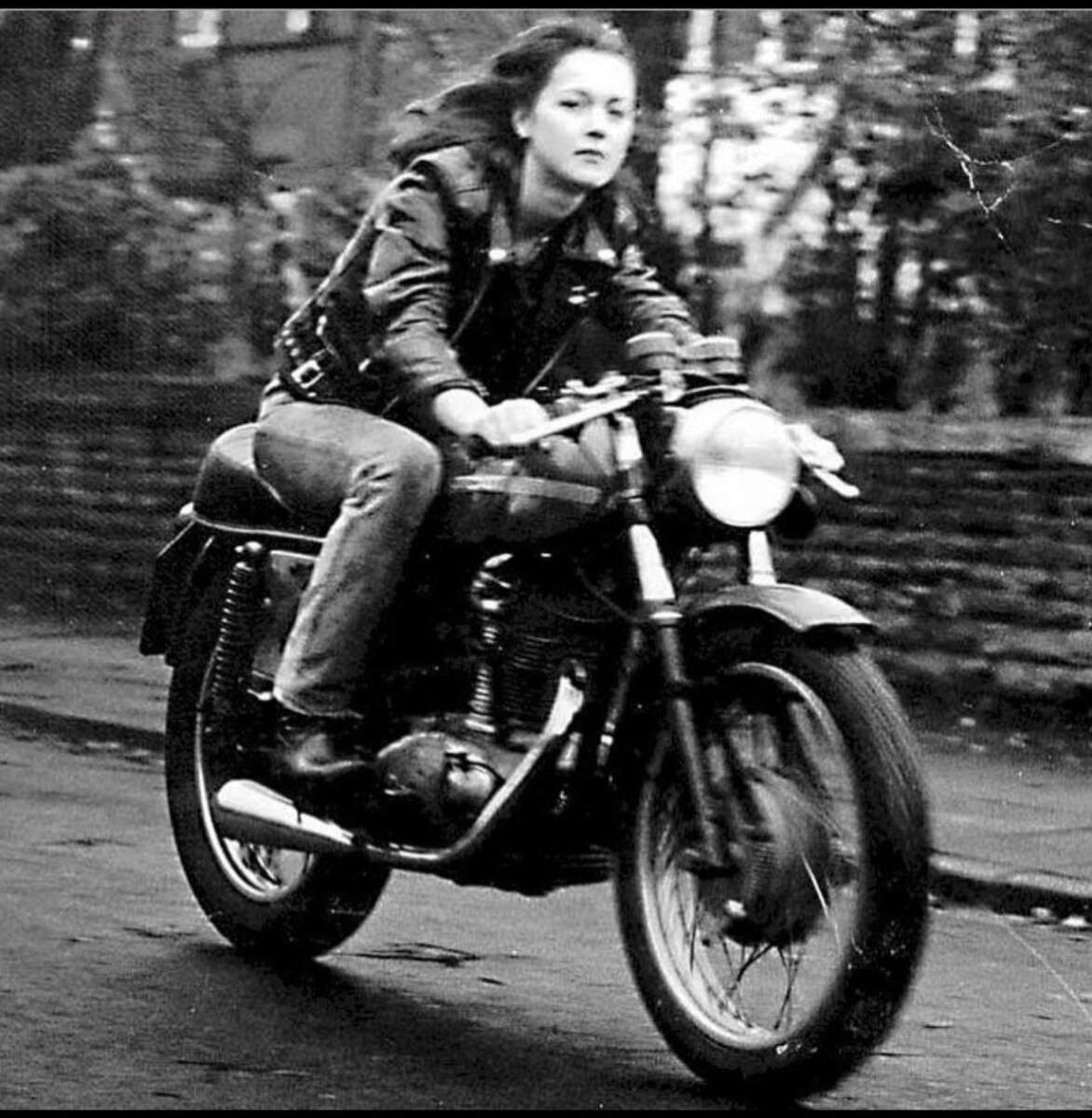 A young woman in a leather jacket rides a vintage motorcycle down a street. The image is black and white, capturing a sense of motion with a blurred background and a focused, determined expression on her face.