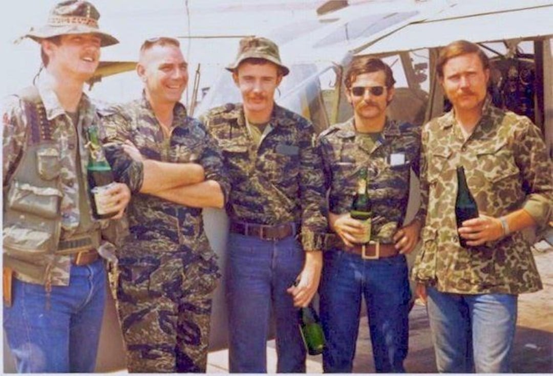 Five men in military uniforms and camouflage clothing stand in front of a helicopter, smiling and holding bottles. The scene appears to be outdoors on a sunny day.