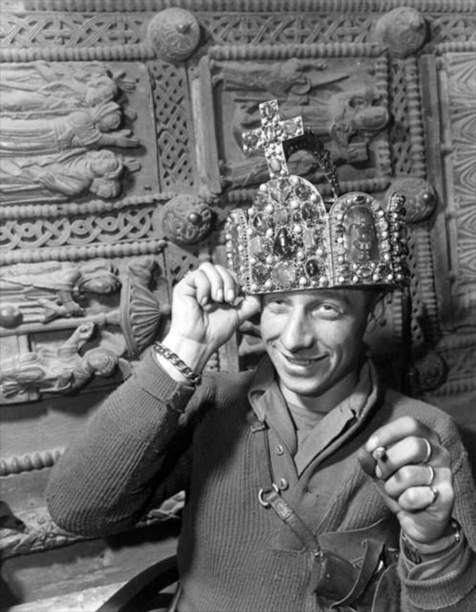 A smiling man in uniform playfully places a large, ornate crown decorated with jewels and a cross on his head, with an intricately carved relief in the background.