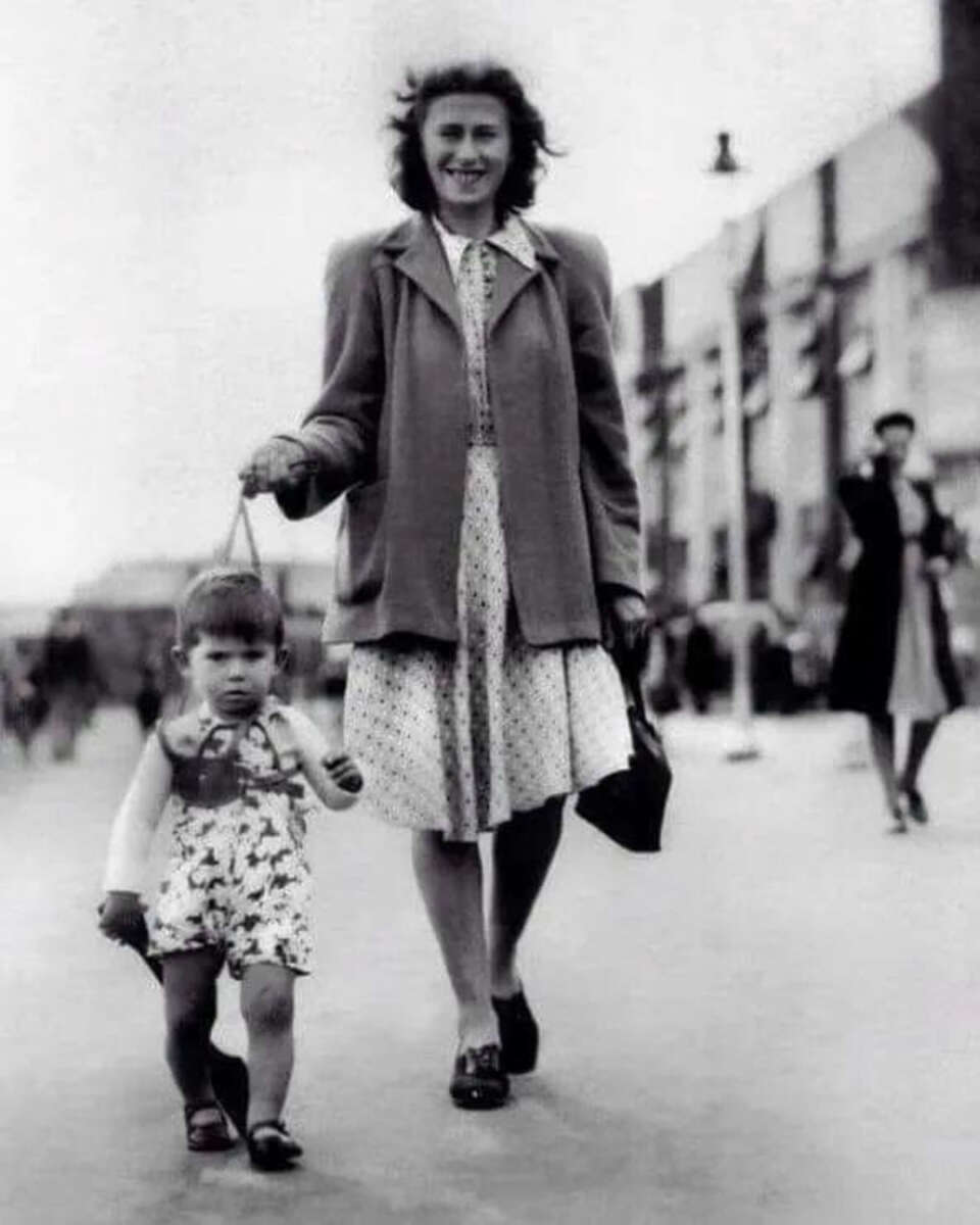 A woman in a dress and coat smiles as she walks outdoors, holding a handbag and leading a small child on a harness. The child looks forward, and people walk in the blurred background. The photo is in black and white.
