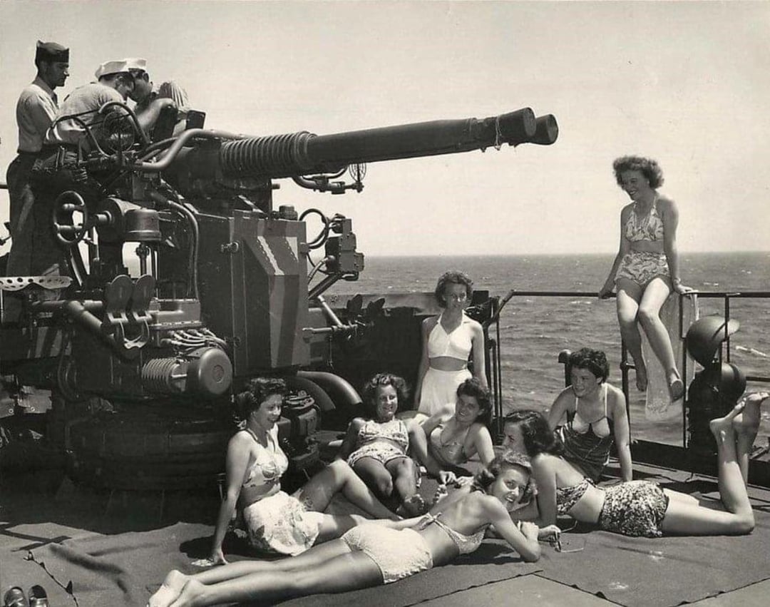 A group of women in vintage swimsuits relax and pose on the deck of a ship near a large naval gun, while two sailors operate the gun in the background; the ocean is visible behind them.