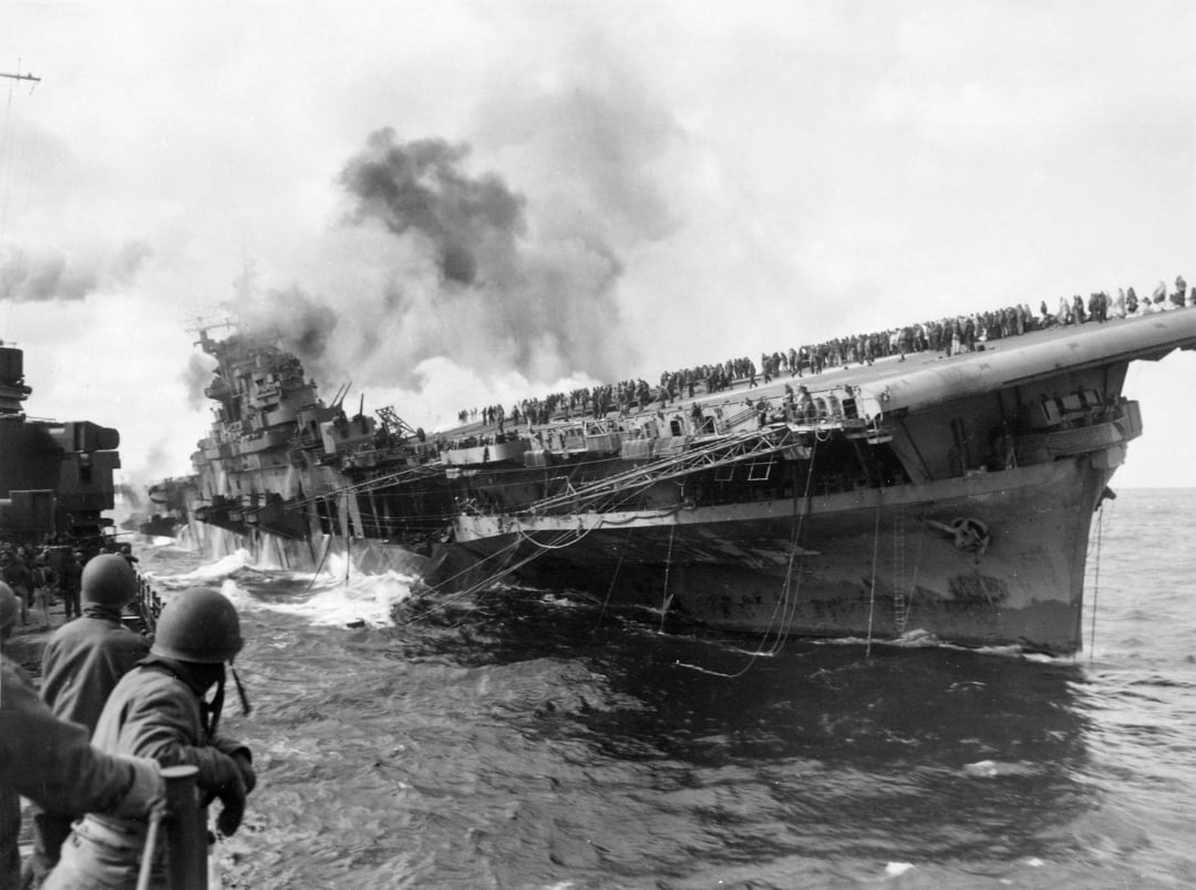 A heavily damaged aircraft carrier listing to one side, with smoke rising and many sailors gathered on the deck, while crew on a nearby ship watch the scene.