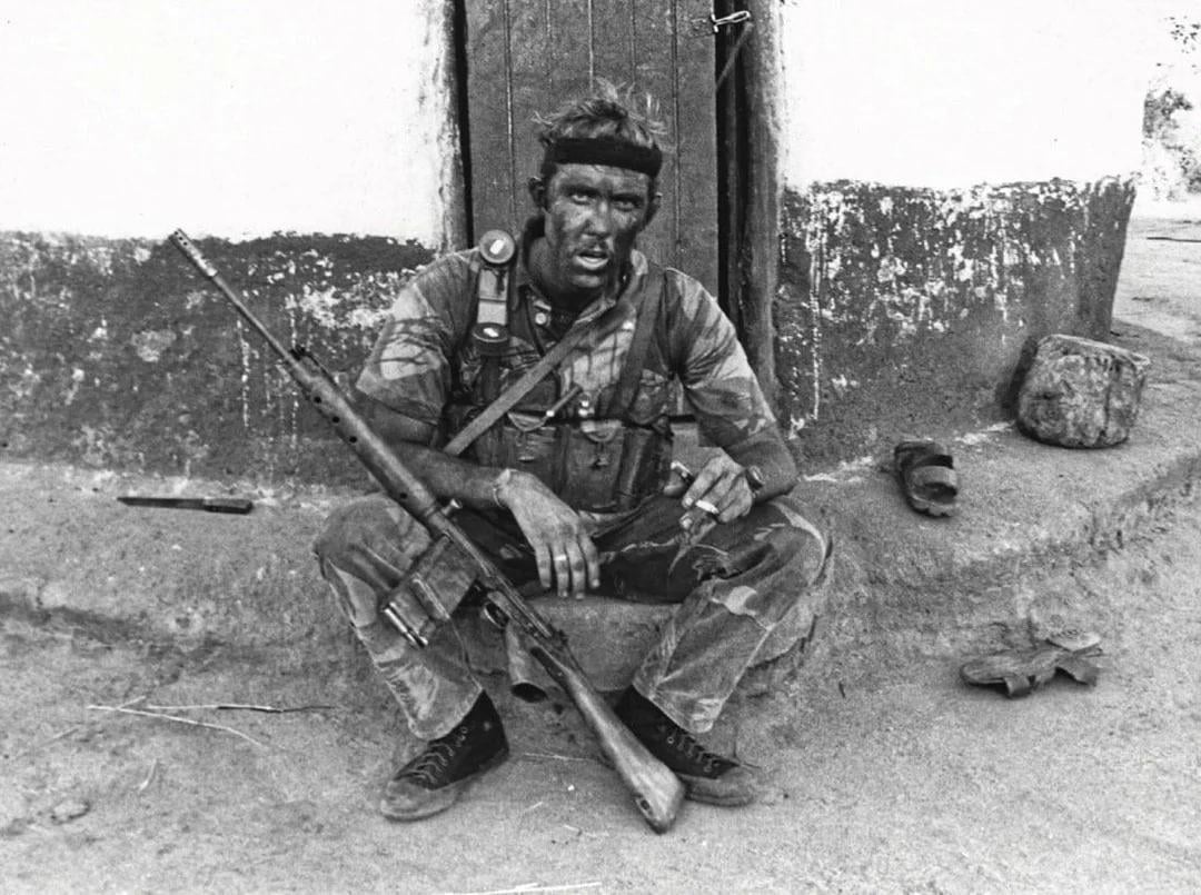 A soldier with a rifle sits on a step outside a worn building, appearing dirty and exhausted. He wears a headband, military gear, and boots, surrounded by equipment and debris on the ground.
