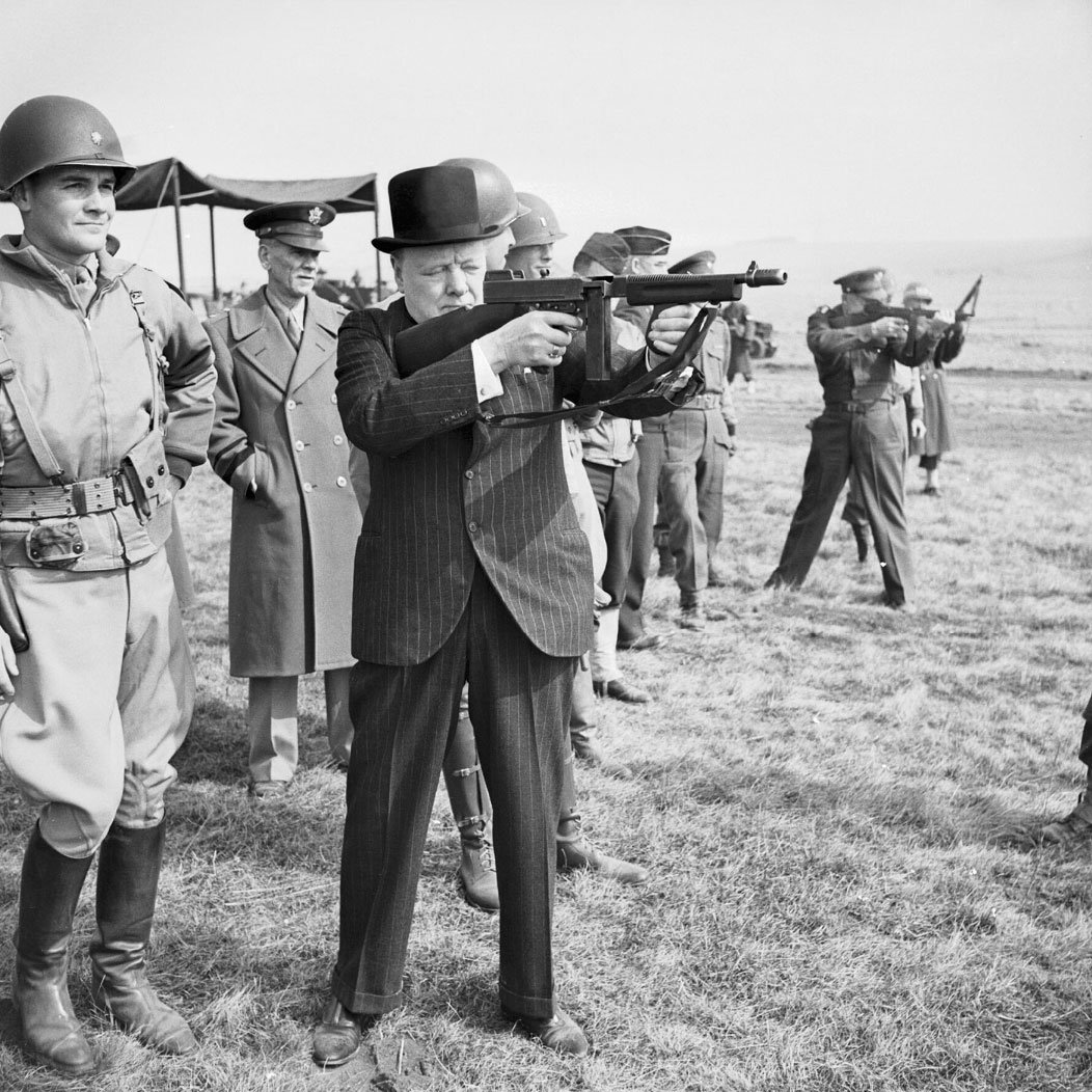 Winston Churchill, wearing a suit and bowler hat, aims a submachine gun at a shooting range, surrounded by uniformed soldiers and military personnel standing on grass.