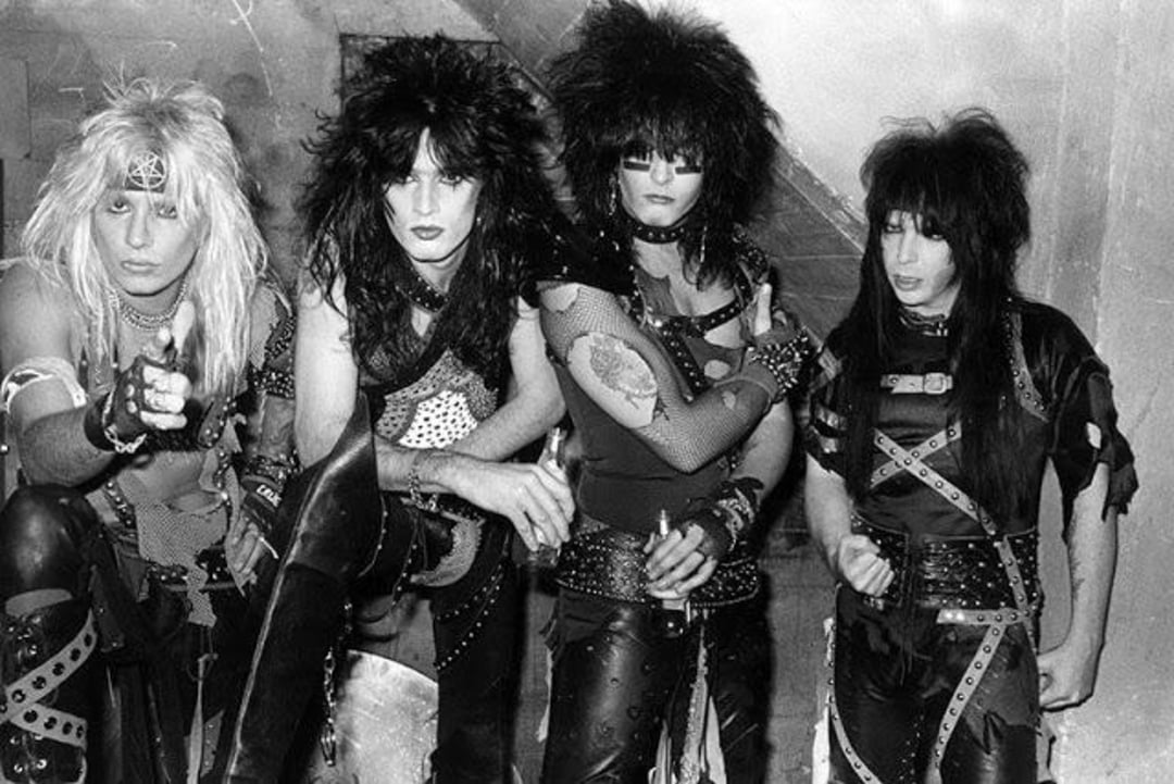 Four men with teased hair and heavy makeup wear black leather outfits with metal studs and accessories, posing against a bare wall in a style typical of 1980s glam metal bands.