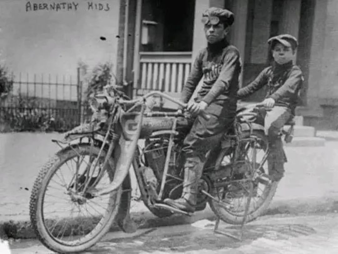 Two young boys, known as the Abernathy kids, sit on an old motorcycle in front of a house. Both wear hats and goggles, and the photo appears to be from the early 1900s.